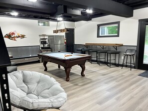 Une table de billard dans une pièce où se trouve un fauteuil en forme de sac de haricots blanc, un coin bar avec des tabourets et un réfrigérateur.