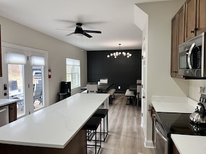 A modern kitchen with a central island, a stove, and a microwave. There are bar stools and a dining area visible.