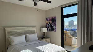 Habitación de hotel con cama, mesita de noche, ventilador de techo, ventana con vista a edificios y un cuadro en la pared.