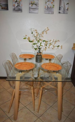 Uma mesa de jantar com tampo de vidro, quatro cadeiras, um vaso com flores e um piso de azulejos.