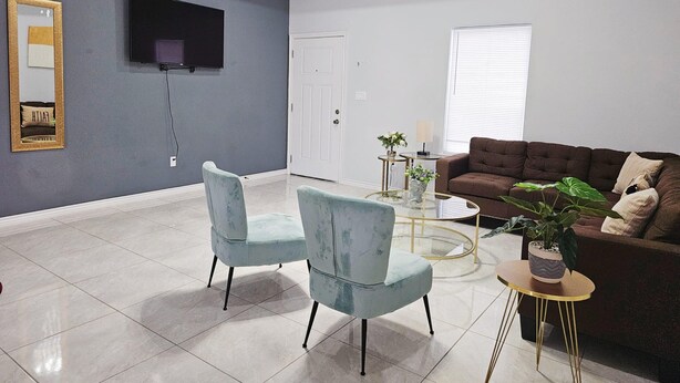 Una sala de estar moderna con un televisor de pantalla plana, dos sillones azul claro, un sofá marrón y una mesa de centro de vidrio.