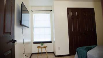 A room with a bed, a door, a window with blinds, a small table with a plant, and a wall-mounted TV.