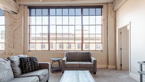 A spacious living room with a large window, a grey sofa, a smaller sofa, and a coffee table. - Beautiful Downtown Condo "Unbeatable Location" with Secure Onsite Parking (New Orleans)