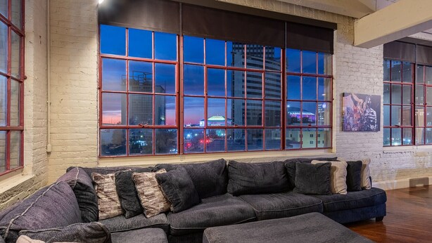 Ein modernes Wohnzimmer mit einer großzügigen Eckcouch, einem großen Fenster mit Blick auf die Stadt und einer Wand mit freiliegenden Ziegelsteinen.