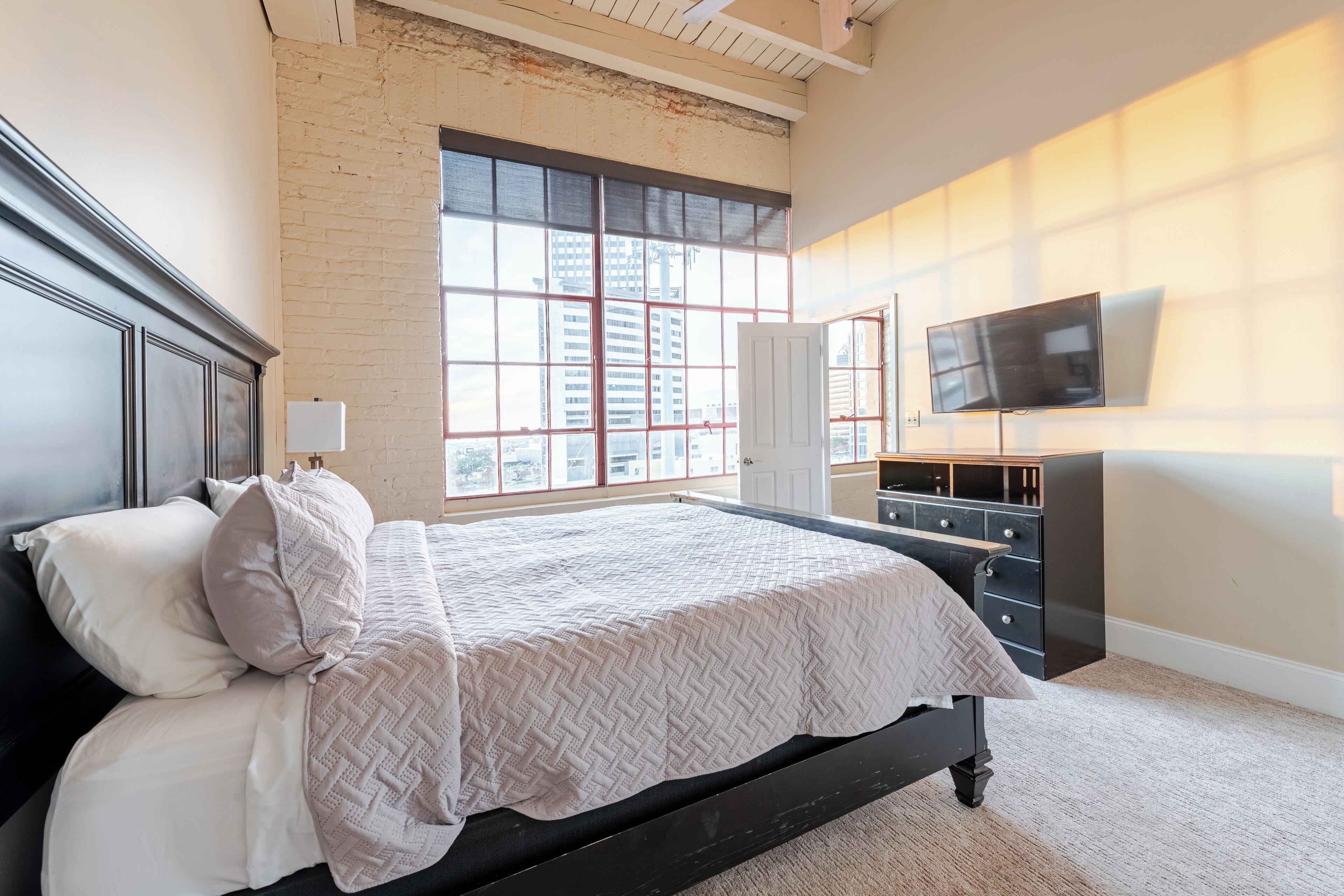A bedroom with a large window, a bed, a dresser, and a TV.