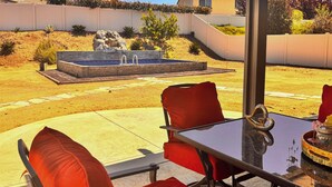 Een terras buiten met een glazen tafel, rode stoelen met kussens en een fonteintje.