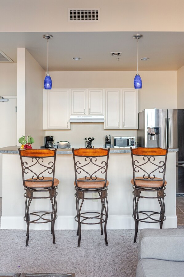A kitchen with three bar stools, a microwave, a refrigerator, and a coffee maker.