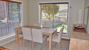 A dining area with a wooden table and chairs, large windows with blinds, and a view of a garden. - Pete's Place. Well-appointed reburnished 2 storey house (South West Rocks)