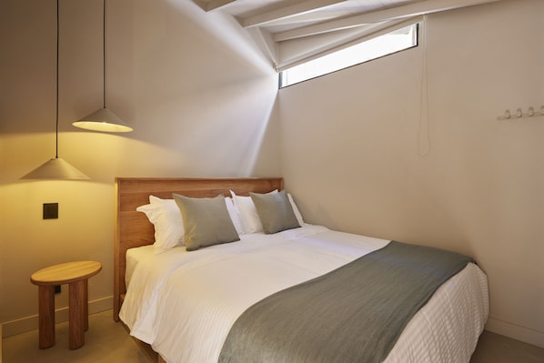 A bedroom with a bed, a wooden headboard, a small stool, a window, and a pendant light. - ANTÍPODA HOTEL (Medellín)