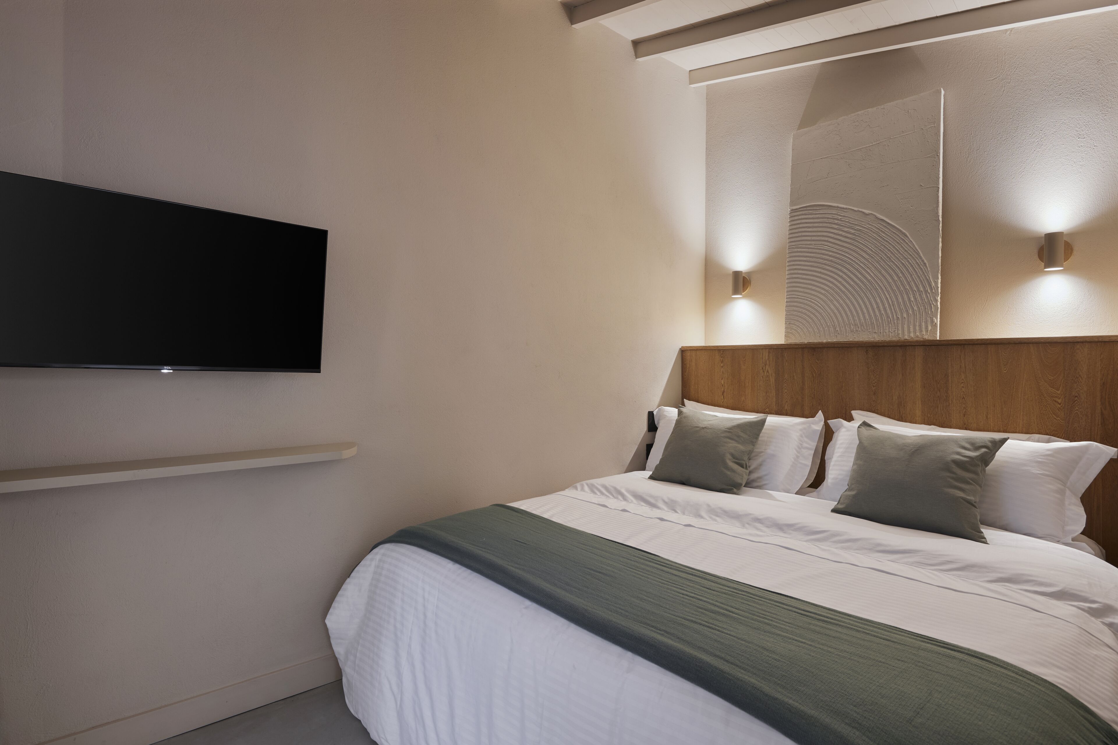 Una cama bien hecha con cabecera de madera, un televisor de pantalla plana montado en la pared y dos lámparas de pared.