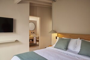Ein Hotelzimmer mit einem Bett, einem an der Wand befestigten Fernseher, einer Nachttischlampe, einem Holz-Kopfteil und einem durch eine offene Tür einsehbarer Badezimmer.
