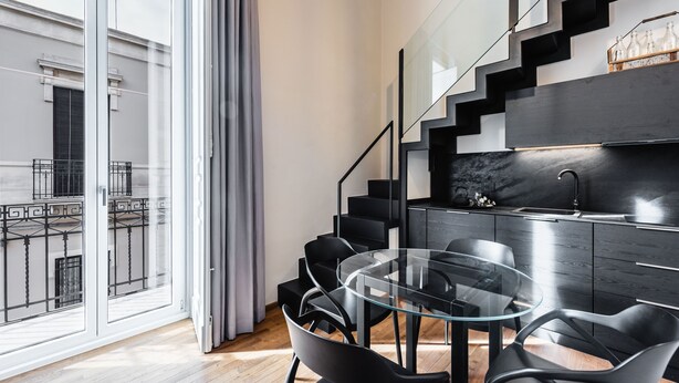 Et moderne køkken med et glasbord og sorte stole, en trappe med gelænder i sort, samt udsigt til en bygning gennem store vinduer.