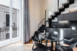 A modern kitchen with a glass dining table and black chairs, a staircase with a black railing, and a view of a building through large windows. - Ferrini Home - Firenze 70 (Catania)