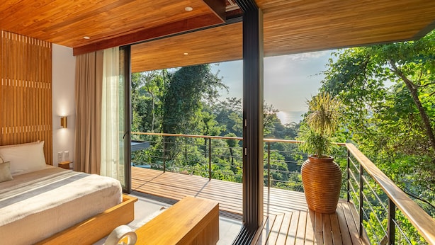 A modern bedroom with a large bed, a wooden bench, and a view of lush greenery through large windows. - Brand-new luxury Villa Element (Provincia de Puntarenas)