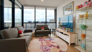 A modern living room with a large window, a flat-screen TV, a sofa with a Pokemon-themed pillow, and a glass cabinet with figurines. - Pokemon Inspired Apt -Pool-Gym-Sauna-Steam Room (Adelaide)
