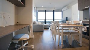 A modern living room with a sofa, dining table, and chairs, a kitchen area with cabinets, and large windows offering a view. - Paradise 2BR Apt in Rundle Mall (Adelaide)