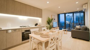 A modern kitchen with a dining area, a grey sofa, and a large window with a city view. - Coastal Retreat in Realm - Luxe 1BR Oasis (Adelaide)