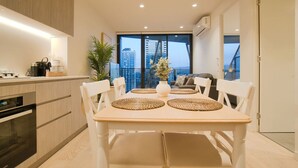 A modern kitchen with a dining area, featuring a large window with a city view, a built-in oven, and a dining table with woven placemats. - Coastal Retreat in Realm - Luxe 1BR Oasis (Adelaide)