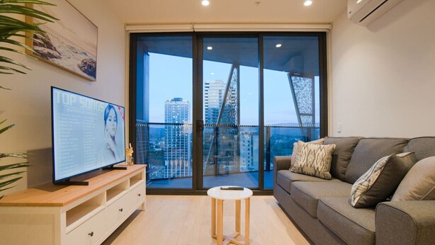 A modern living room with a large flat-screen TV, a grey sofa with patterned cushions, a small round wooden stool, and a large window offering a city view. - Coastal Retreat in Realm - Luxe 1BR Oasis (Adelaide)