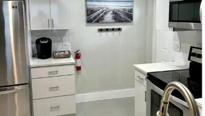 Cozinha compacta com eletrodomésticos de aço inoxidável, bancada branca e extintor de incêndio na parede.