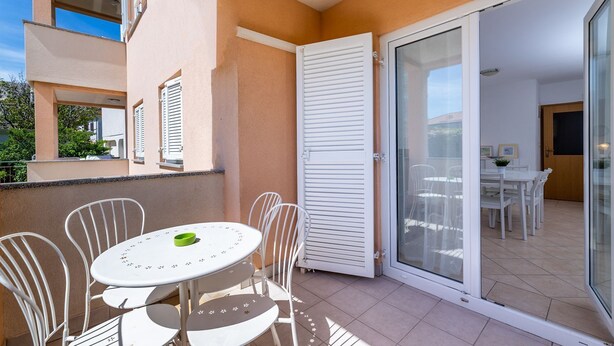 Un balcone con tavolo e sedie, una porta scorrevole in vetro che dà su un'area pranzo interna, e la facciata di un edificio con persiane.