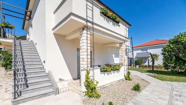 A modern two-story building with a stone facade, a covered outdoor area with a staircase, and a garden with small plants and flowers. - Only 300 metres to the great beach (Baska)