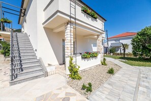 Un bâtiment moderne à deux étages, doté d’une façade en pierre, d’un espace extérieur couvert avec un escalier, et d’un jardin agrémenté de petites plantes et de fleurs.