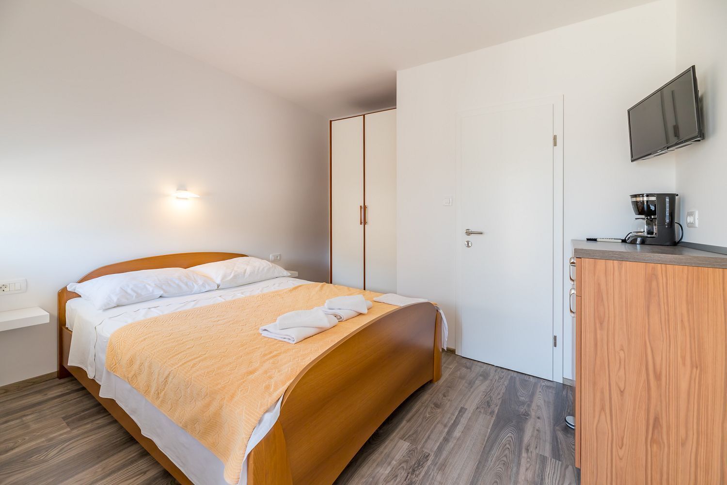 A neatly made bed with a wooden headboard, a bedside table with a coffee maker, and a wardrobe with sliding doors.