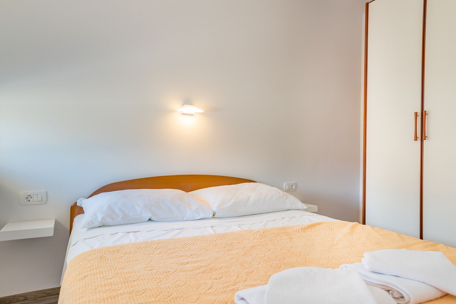 Un lit bien fait, avec du linge de lit blanc et un plaid jaune, une table de chevet avec une lampe, et une armoire avec des poignées en bois.