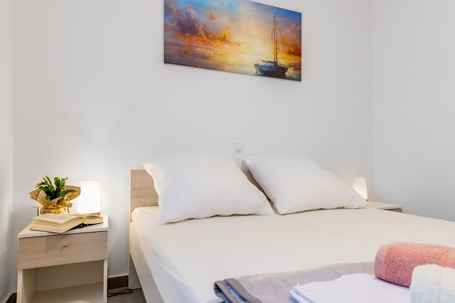 A bedroom with a bed, a bedside table with a lamp, and a painting of a sailboat on the wall.