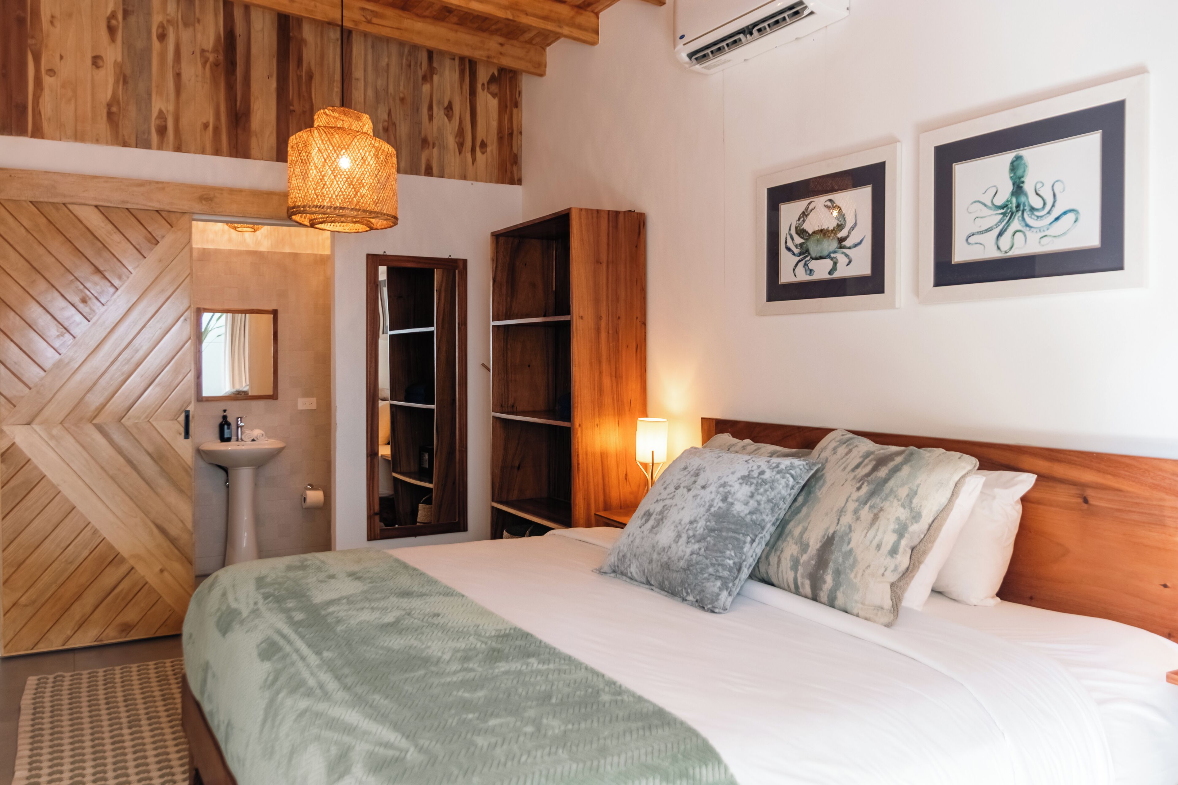 Un dormitorio con cama, techo de madera, estante en la pared, mesita de noche con lámpara y cuadros enmarcados.