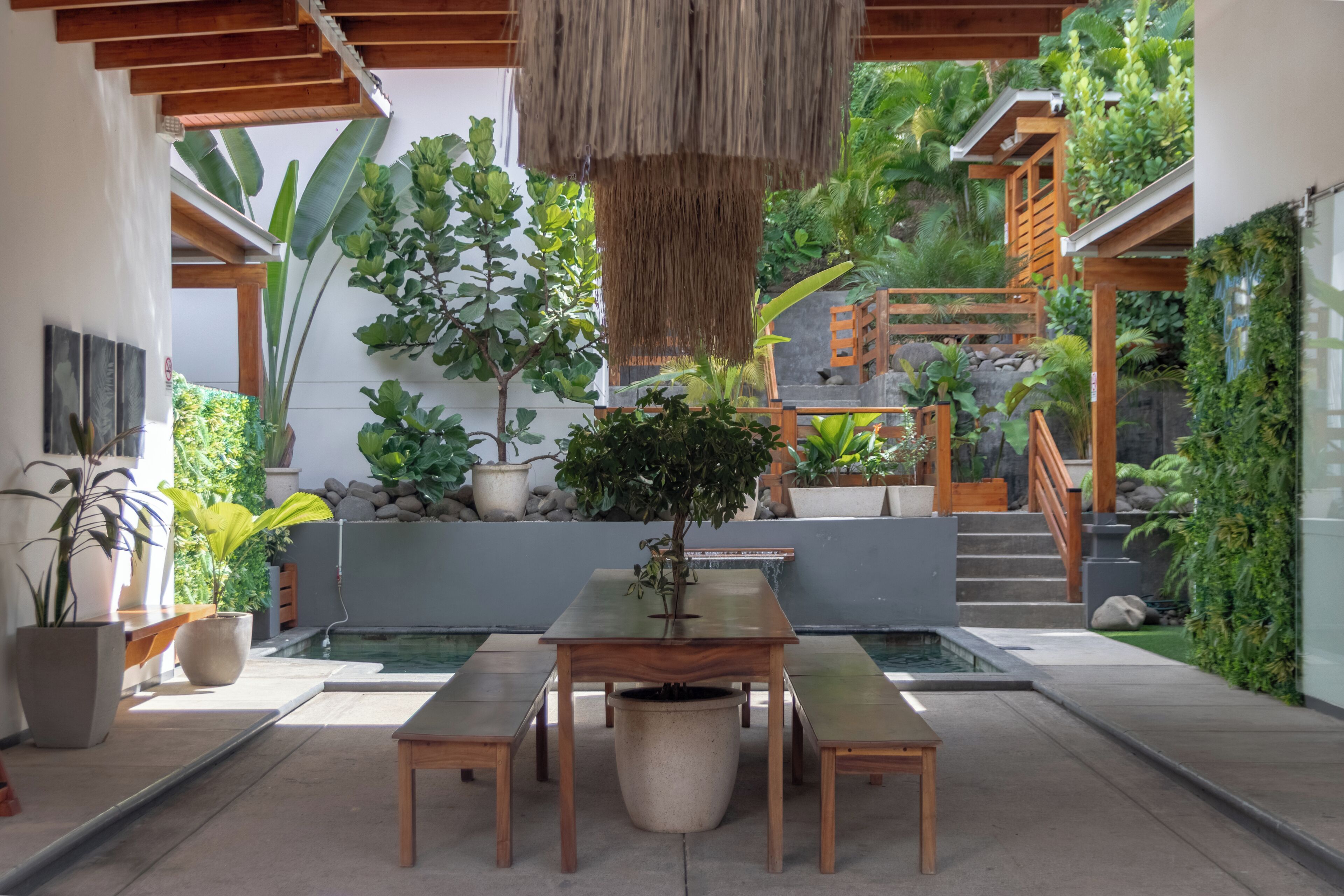 A modern outdoor dining area with a wooden table and benches, surrounded by potted plants and a small water feature.
