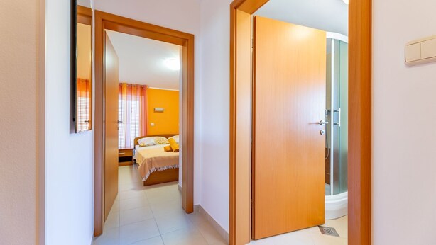 Un couloir mène à une chambre à coucher avec un lit, une salle de bain avec une douche et une porte visible.