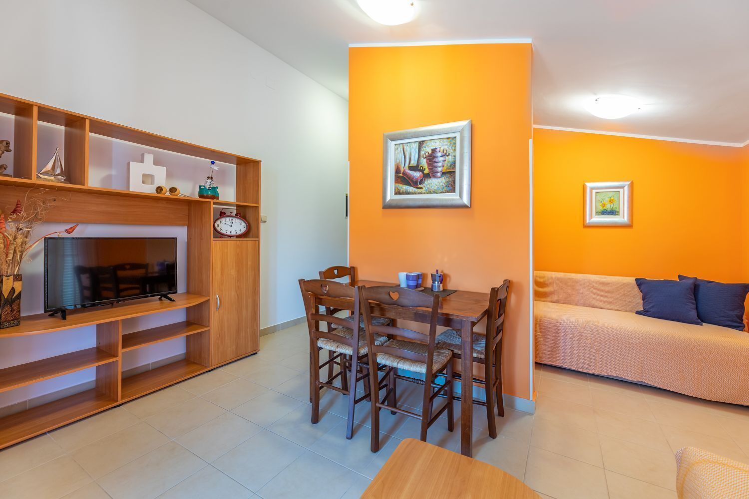 Un salon avec un téléviseur, des étagères en bois, une table à manger avec des chaises, un mur orange et un canapé.