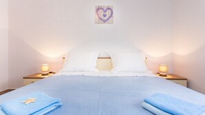 A neatly made bed with white pillows and a blue bedspread, two bedside lamps, and a heart-shaped decoration on the wall. - 200 metres to the crystal clear sea (Baska)