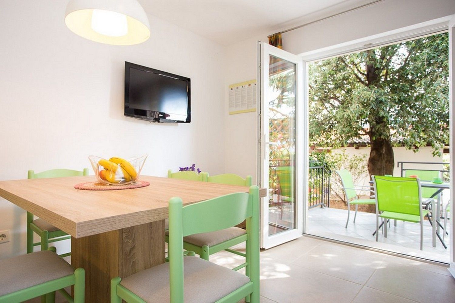 A dining area with a wooden table, green chairs, a fruit bowl, and a TV mounted on the wall.