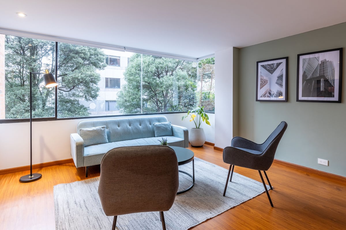 A modern living room with a sofa, a chair, a coffee table, and a large window with a view of trees and buildings.