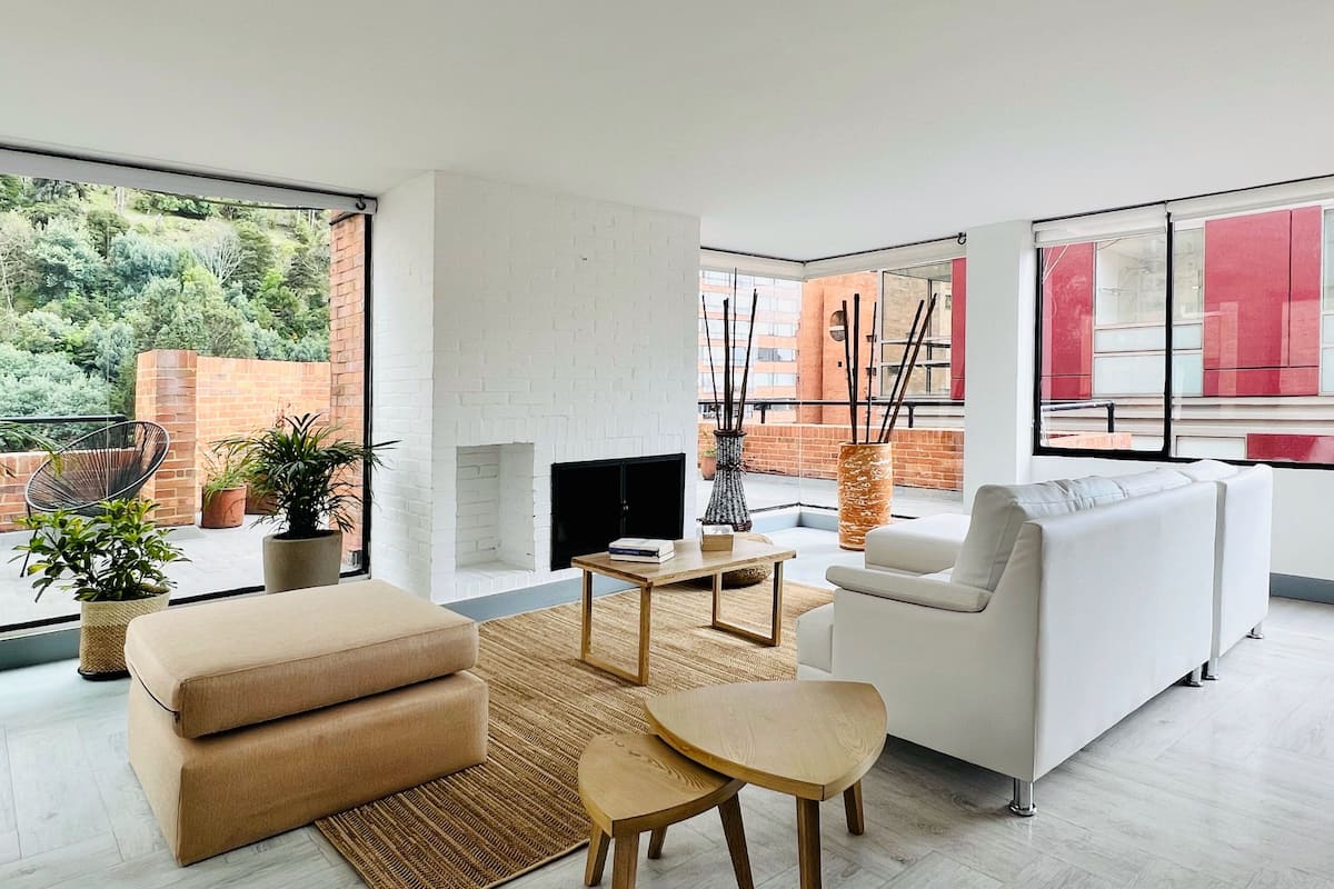 A modern living room with a white sofa, a beige couch, a small wooden table, and a fireplace. There are potted plants and a view of a brick wall and greenery outside.