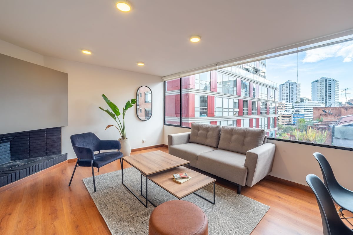 A modern living room with a sofa, a coffee table, and a chair. There is a large window with a city view.