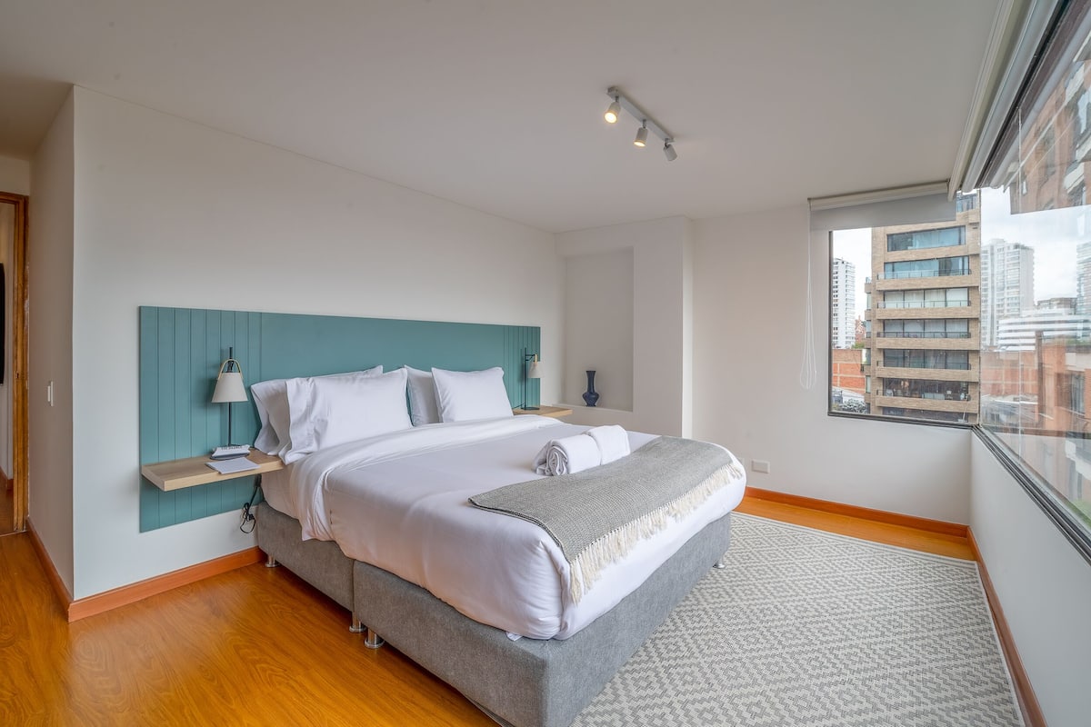 A modern bedroom with a large bed, a teal headboard, a bedside table with a lamp, and a view of the city through a window.