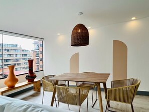 A modern dining area with a wooden table and wicker chairs, a hanging light fixture, and a view of a cityscape through a large window. - Chicala By Wynwood House (Bogotá)