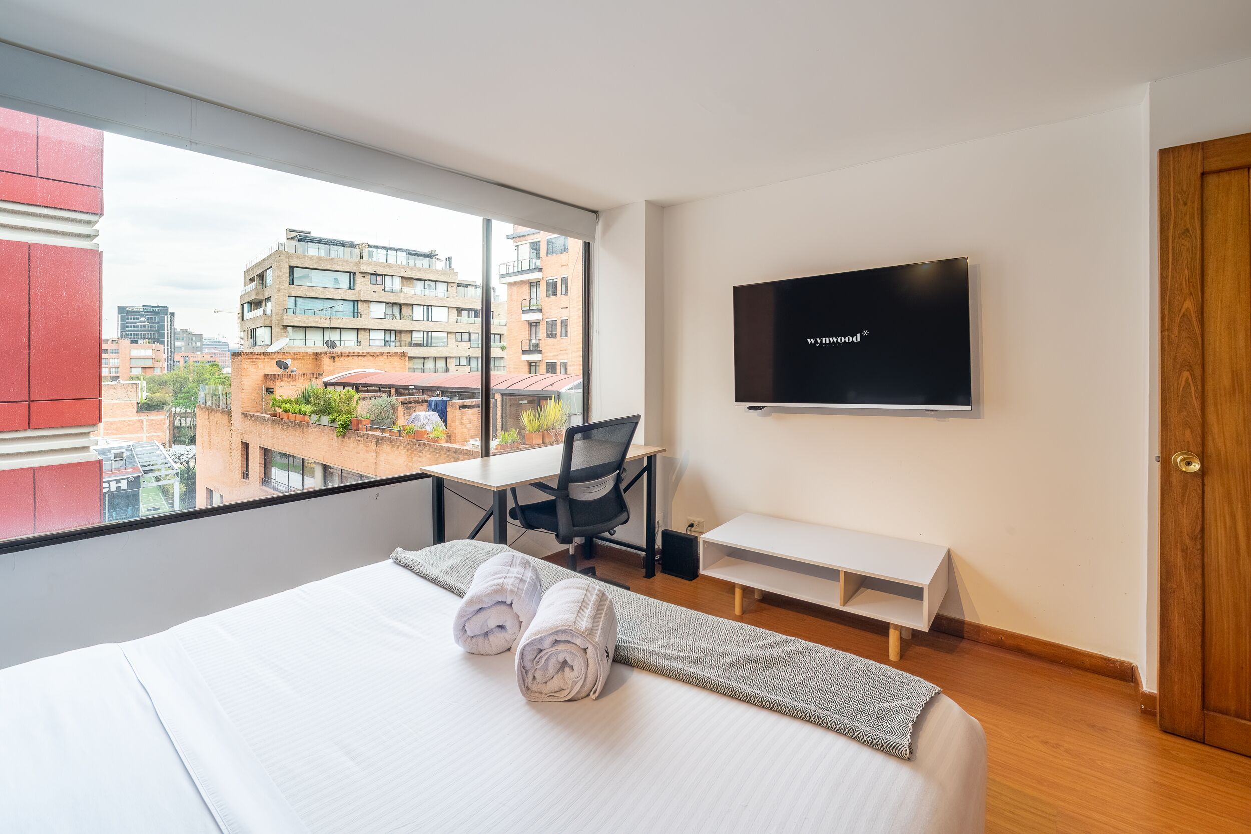 A hotel room with a bed, a desk, a chair, a TV, and a view of buildings outside.