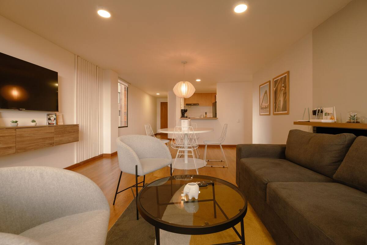 A modern living room with a glass coffee table, grey sofa, and a dining area with a white table and chairs.