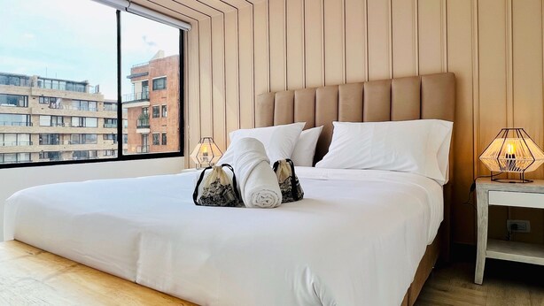 A modern hotel room with a large bed, wooden headboard, and a view of buildings through a large window. - Chicala By Wynwood House (Bogotá)