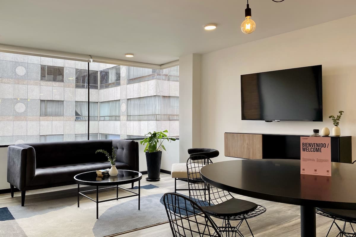 A modern living room with a black sofa, a round coffee table, and a dining table with chairs. A large TV is mounted on the wall, and a welcome sign is visible.