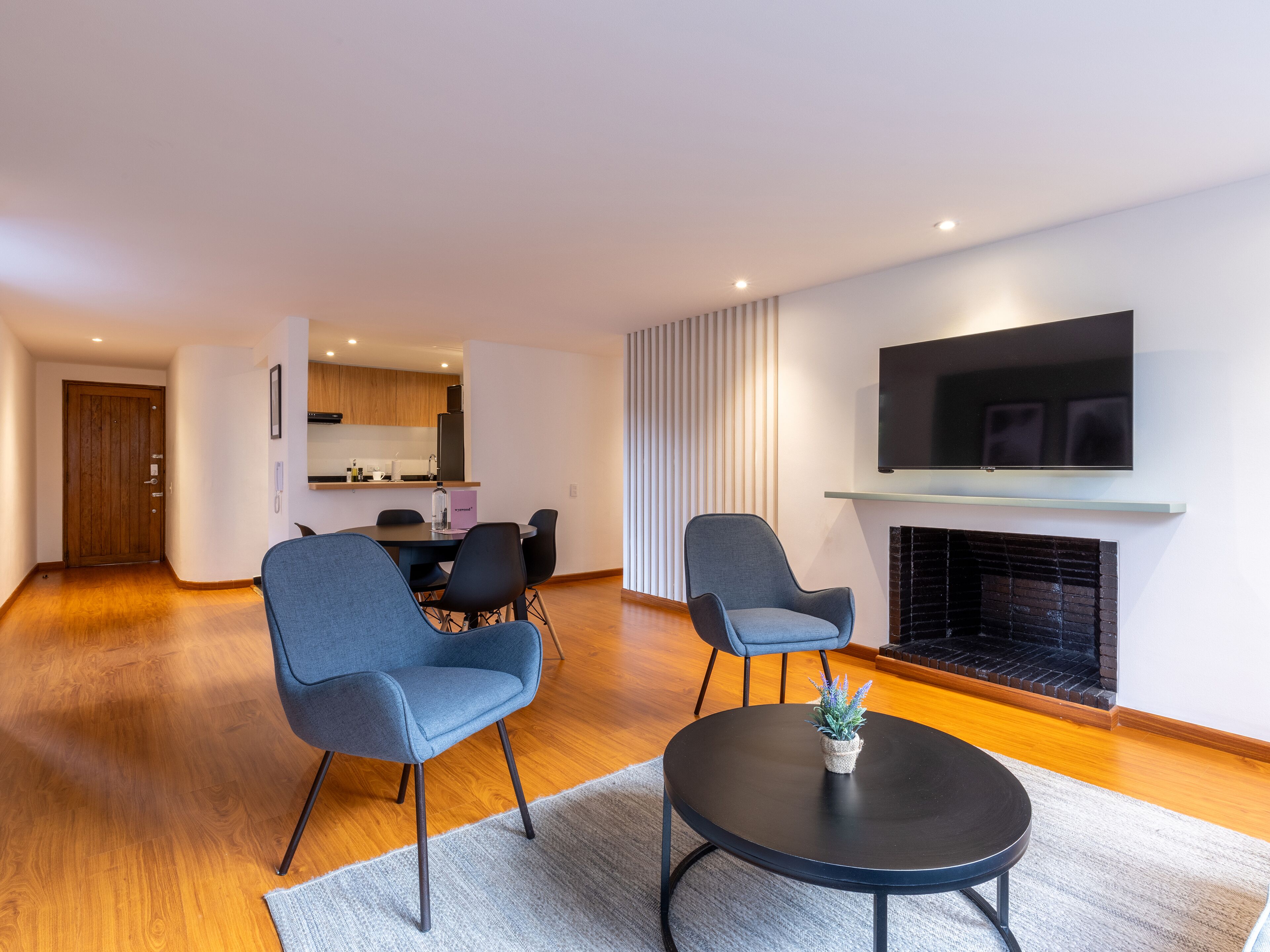 A modern living room with a round black coffee table, two blue armchairs, a flat-screen TV mounted above a fireplace, and a wooden floor.