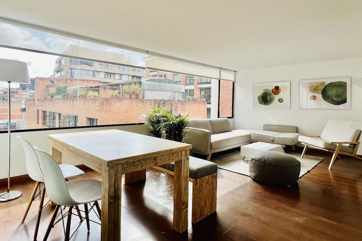 A modern living room with a large window, a wooden dining table, a sofa, and a coffee table.