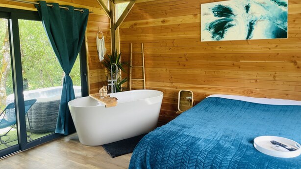 A wooden cabin room with a large window, a bathtub, a bed, and a painting on the wall.