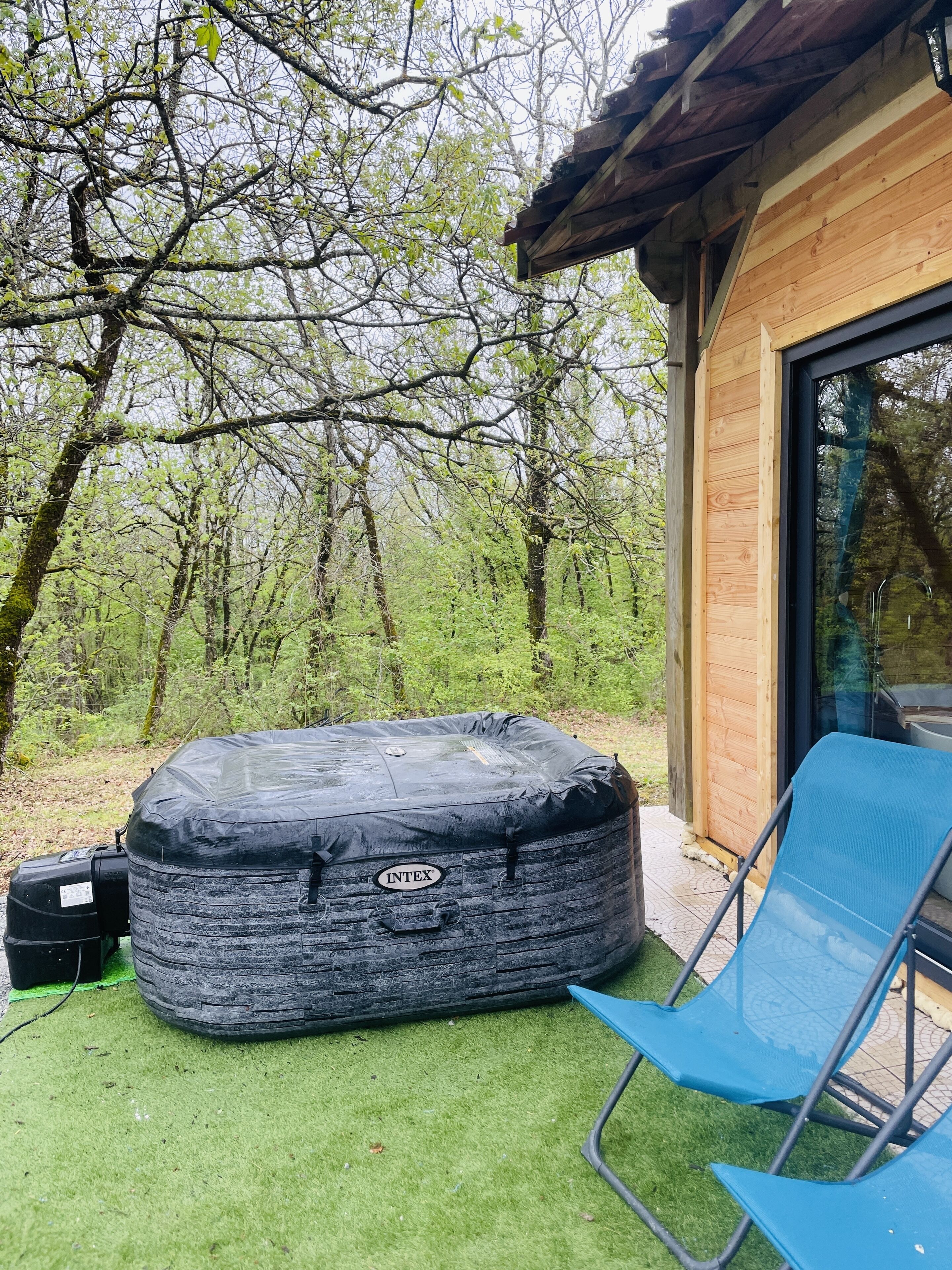 Un jacuzzi con una bomba, una silla azul y una cabaña de madera con puerta de vidrio.