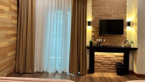 Una habitación moderna con suelo de madera, un ventanal con cortinas translúcidas, una pared de ladrillo decorativa, un televisor montado y una pequeña mesa con un cubo de basura.